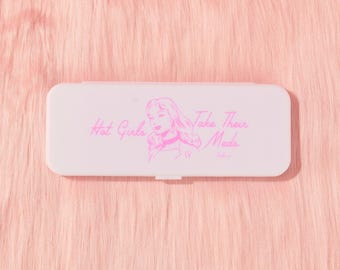 Puede incluir: Un pastillero rectangular de color rosa claro con esquinas redondeadas. La superficie superior presenta un dibujo lineal de color rosa intenso de una persona con pelo largo y el texto Hot Girls Take Their Meds en cursiva.