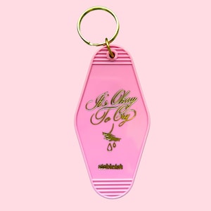 May include: Pink motel-style keychain with gold lettering that reads "It's Okay To Cry" and a weeping eye graphic. The keychain has a gold ring and the word "stebielish" at the bottom.