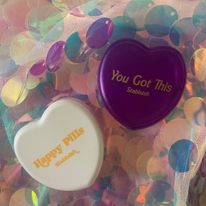 May include: Two heart-shaped pill boxes, one purple and one white, with the text "You Got This" and "Happy Pills" in gold lettering. Both boxes have the word "Stableish" printed below the text.