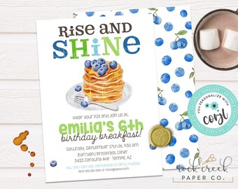 Puede incluir: Una invitación blanca con texto azul y verde que dice "Rise and Shine" y presenta una ilustración de acuarela de una pila de panqueques con arándanos y jarabe. La invitación es para el desayuno de cumpleaños de Emilia por sus 6 años en el Barnyard Breakfast Diner. La invitación incluye la fecha, la hora, la dirección y la información de RSVP.
