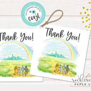 May include: Two printable thank you tags featuring a watercolor illustration of the yellow brick road from the Wizard of Oz. The illustration includes the characters Dorothy, the Scarecrow, the Tin Man, and the Cowardly Lion walking down the road towards the Emerald City. A rainbow arches over the city. The text "Thank You!" is printed on each tag.