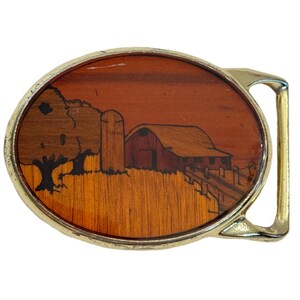 May include: Oval belt buckle with a gold-tone frame. The buckle features a wooden inlay depicting a rural scene with a barn, silo, tree, and fence. The wood has various shades of brown and orange.