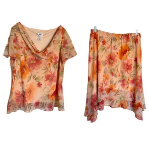 May include: A two-piece floral outfit featuring a short-sleeved top and a skirt. The top has a cowl neck and the skirt has a flowing, asymmetrical hem. The fabric is a sheer peach color with a floral pattern in shades of orange, pink, and green.