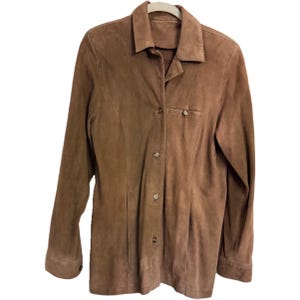 May include: A brown suede button-up shirt with long sleeves. The shirt has a collared neckline, a single chest pocket, and decorative stitching. The shirt is hanging on a white hanger against a white background.