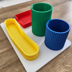 May include: A white desk organizer base featuring a long yellow tray, a square red tray, a tall green cylindrical cup, and a shorter blue cylindrical cup. Each component has a ribbed, textured exterior surface and a solid, flat interior.