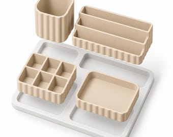 Puede incluir: Un conjunto organizador de escritorio beige que incluye un vaso cilíndrico alto, una bandeja rectangular escalonada, una bandeja cuadrada de seis compartimentos y una bandeja rectangular, sobre una base blanca con secciones.