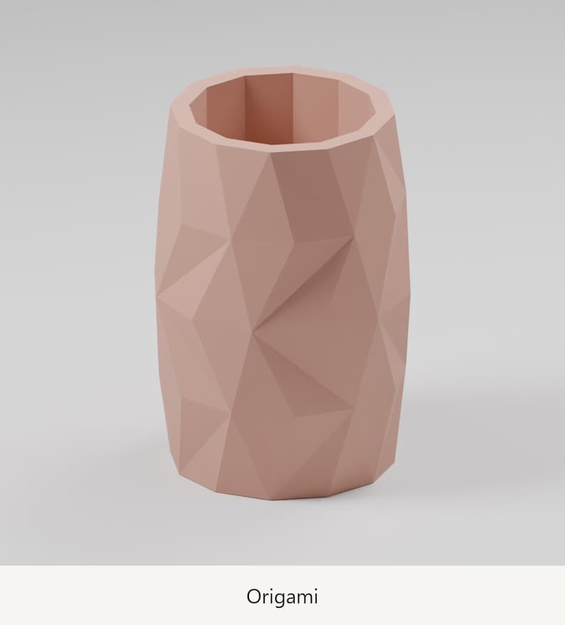 May include: A dusty rose cylindrical vase featuring a faceted, geometric exterior surface with sharp, angular planes. The vessel has an open top and a hollow interior, displaying a consistent matte finish throughout its entire structure.
