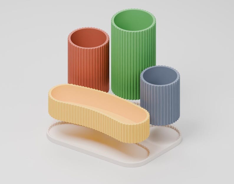 Puede incluir: Un conjunto organizador de escritorio con tres recipientes cil&iacute;ndricos acanalados en terracota, verde y azul, junto a una bandeja ovalada alargada acanalada en amarillo claro, sobre una base blanca con un borde acanalado a juego.