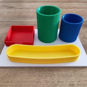 Puede incluir: Un organizador de escritorio blanco con una bandeja cuadrada roja, un vaso cil&iacute;ndrico verde alto, un vaso cil&iacute;ndrico azul m&aacute;s corto y una bandeja ovalada amarilla larga, todos con una textura exterior acanalada.