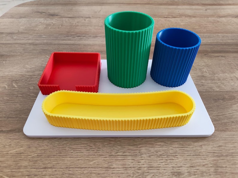 Puede incluir: Un organizador de escritorio blanco con una bandeja cuadrada roja, un vaso cil&iacute;ndrico verde alto, un vaso cil&iacute;ndrico azul m&aacute;s corto y una bandeja ovalada amarilla larga, todos con una textura exterior acanalada.