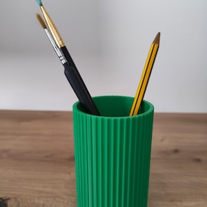 May include: A bright green cylindrical pencil holder with a vertical ribbed texture. The container holds two paintbrushes with black handles and one yellow and black striped pencil, all standing upright inside the open top.