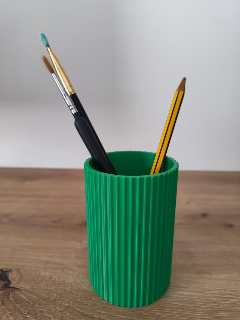 May include: A bright green cylindrical pencil holder with a vertical ribbed texture. The container holds two paintbrushes with black handles and one yellow and black striped pencil, all standing upright inside the open top.