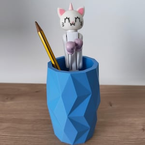 May include: A blue geometric pencil holder containing a yellow pencil and a white cat-unicorn figure with pink ears and purple boxing gloves. The cat-unicorn has closed eyes, a small horn, and a white body, standing upright inside the faceted container.