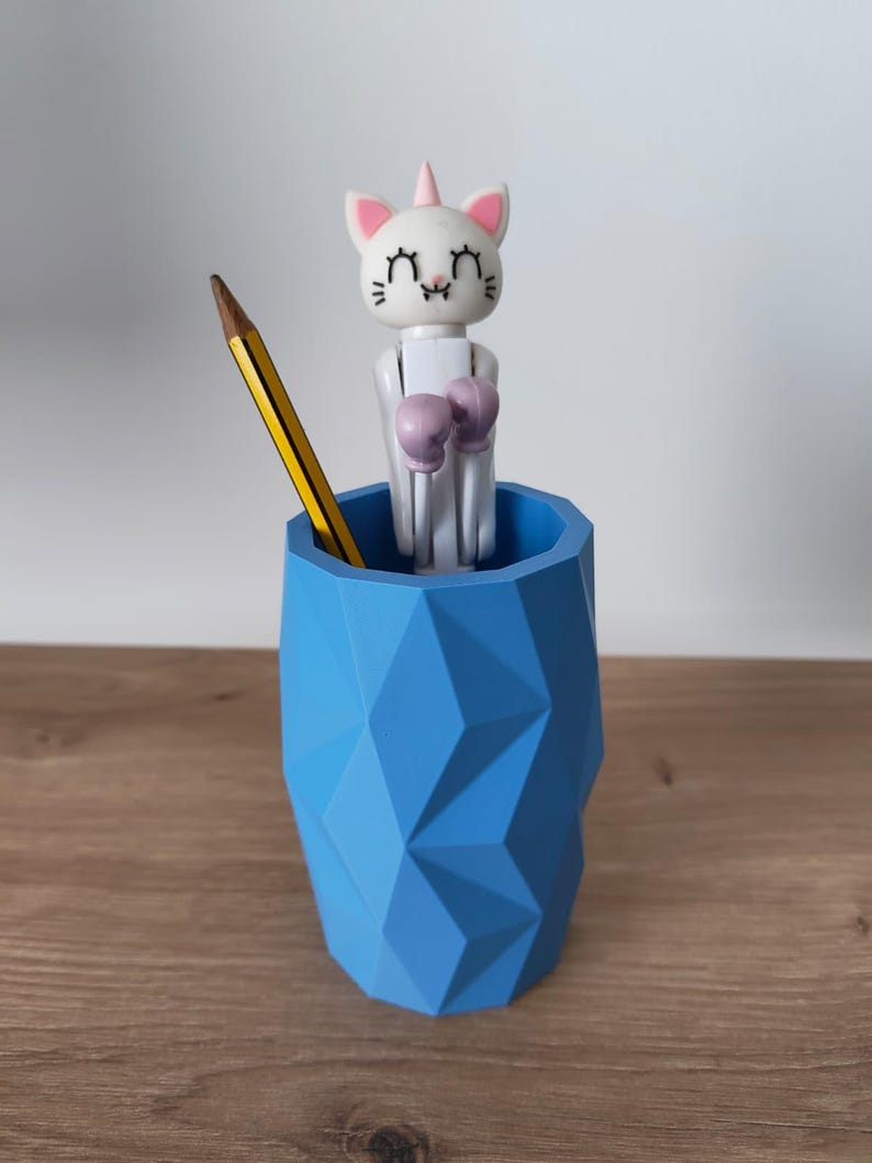 May include: A blue geometric pencil holder containing a yellow pencil and a white cat-unicorn figure with pink ears and purple boxing gloves. The cat-unicorn has closed eyes, a small horn, and a white body, standing upright inside the faceted container.