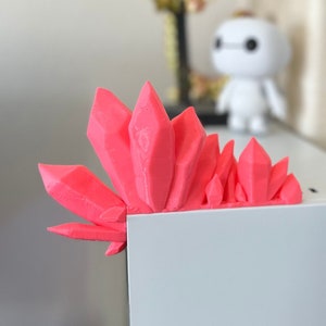 May include: A bright pink 3D printed crystal-shaped object sits on the edge of a white surface.