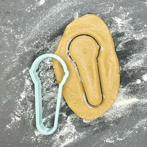 Pode incluir: Um cortador de biscoitos de plástico azul claro em forma de cone de sorvete, ao lado de uma massa de biscoitos marrom com a forma de cone de sorvete recortada.