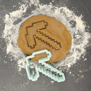 May include: A light blue plastic cookie cutter shaped like a Minecraft pickaxe. The cutter is on top of a brown cookie dough circle.