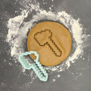 May include: A light blue plastic cookie cutter shaped like a pickaxe on a brown cookie dough background.