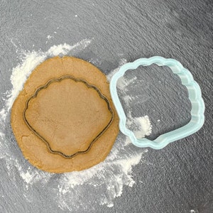May include: A light blue plastic seashell cookie cutter on a grey surface with a brown cookie dough circle underneath it. The cookie cutter is on top of the dough and is ready to be pressed into the dough.