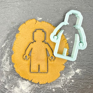 May include: A light blue plastic cookie cutter shaped like a Lego person on a brown cookie dough background.