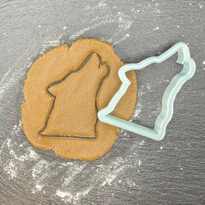Puede incluir: Un cortador de galletas de lobo de plástico azul claro sobre un círculo de masa de galleta marrón. El cortador de galletas tiene la forma de un lobo aullando a la luna.