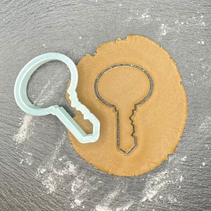 Op de afbeelding: Een lichtblauwe plastic koekjesvormer in de vorm van een sleutel, op een ronde bruine koekdeeg.