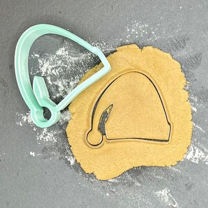 Puede incluir: Un cortador de galletas de plástico verde con forma de gorro de Papá Noel se encuentra sobre una superficie gris junto a una masa de galletas marrón con la forma del gorro de Papá Noel recortada.