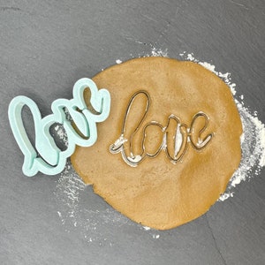 May include: A light blue plastic cookie cutter in the shape of the word "love" is placed on top of a brown cookie dough circle. The cookie cutter is used to cut out the word "love" from the dough.