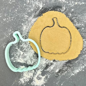 Może przedstawiać: Niebieska plastikowa foremka do ciasteczek w kształcie dyni na szarym tle z wyciętym brązowym kółkiem ciasta.