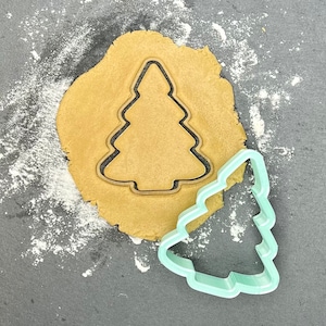 May include: A green plastic Christmas tree cookie cutter on a gray surface with a brown cookie dough circle cut out with the same shape.