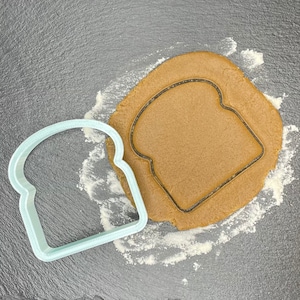 以下が含まれることがあります： グレーの表面に、パンの輪切り形をした薄い青色のプラスチック製のクッキーカッターが置かれ、その中に茶色のクッキー生地の切り抜きがある。