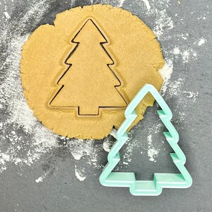 May include: A green plastic Christmas tree cookie cutter on a surface covered in white flour, next to a round piece of cookie dough with a Christmas tree shape cut out of it.