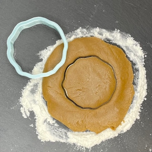 May include: A light blue plastic cookie cutter in the shape of a circle with scalloped edges is placed on top of a round piece of brown cookie dough. The dough is on a black surface covered in white flour.