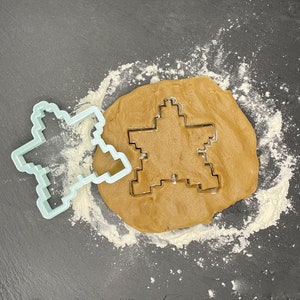May include: A light blue plastic cookie cutter in the shape of a star is placed on top of a brown gingerbread cookie dough. The cookie dough is on a gray surface covered in white flour.