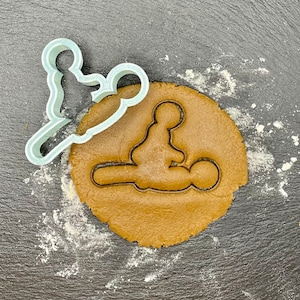 May include: A light blue plastic cookie cutter in the shape of a person sitting down. The cookie cutter is on top of a brown gingerbread cookie dough.