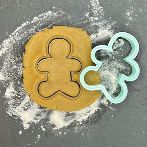 May include: A gingerbread man cookie cutter on a surface dusted with flour, next to a light blue gingerbread man cookie cutter.