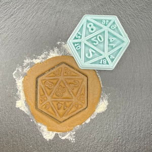 May include: A light blue plastic cookie cutter shaped like a twenty-sided die with numbers 4, 6, 8, 10, 12, 14, 16, 18, 20, and the number 8 repeated twice. The cookie cutter is on a gray surface with a brown cookie dough pressed into the cutter.