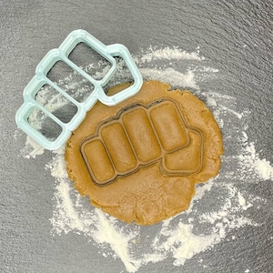 May include: A light blue plastic cookie cutter shaped like a fist with four knuckles. The cookie cutter is on top of a brown cookie dough that has been pressed into the shape of the cookie cutter.