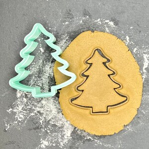 May include: A green plastic Christmas tree cookie cutter on top of a brown cookie dough circle. The cookie cutter is being used to cut out the shape of a Christmas tree in the dough.