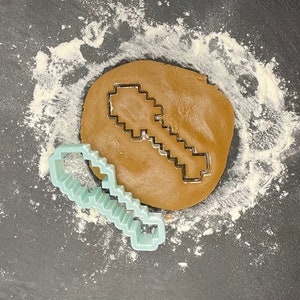 May include: A light blue plastic cookie cutter shaped like a Minecraft pickaxe. The cutter is on top of a brown cookie dough circle.