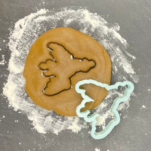 Peut inclure: Un emporte-pièce en forme d'oiseau bleu clair est posé sur une surface grise avec un cercle de pâte à biscuits brune derrière lui. L'emporte-pièce est partiellement sur la pâte.