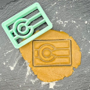 Puede incluir: Un cortador de galletas de plástico verde en forma de círculo estilizado con tres líneas horizontales. El cortador de galletas está encima de una masa de galletas marrón con el mismo diseño impreso en ella.