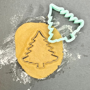 May include: A green plastic Christmas tree cookie cutter on top of a round piece of brown cookie dough. The cookie cutter is being used to cut out the shape of a Christmas tree.