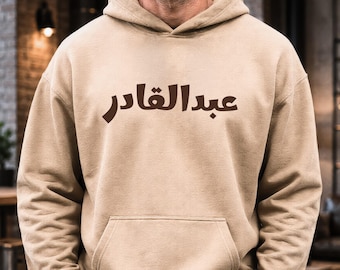 May include: A beige hooded sweatshirt featuring a front kangaroo pocket and dark brown Arabic text printed across the chest. The garment has long sleeves, a drawstring-free hood, and a solid, neutral-toned fabric construction.