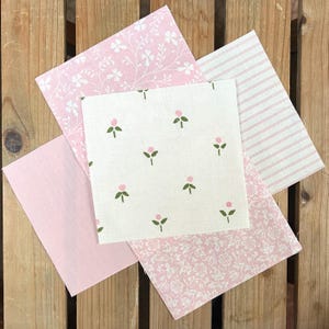May include: Five pink fabric squares arranged on a wooden surface. The fabrics feature various patterns, including floral, striped, and solid designs. The color palette is soft, with shades of pink and white, creating a delicate and feminine aesthetic.