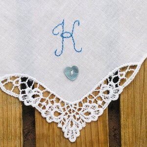Peut inclure: Un mouchoir blanc avec une lettre H brodée en bleu et un bouton bleu en forme de cœur. Le mouchoir est bordé d'une dentelle blanche.