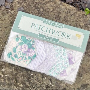 Pode incluir: Um pacote de pe&ccedil;as de patchwork Laura Ashley. O pacote cont&eacute;m v&aacute;rios peda&ccedil;os de tecido com desenhos florais e estampados em tons de branco, roxo e verde-azulado. O pacote &eacute; selado em pl&aacute;stico transparente.