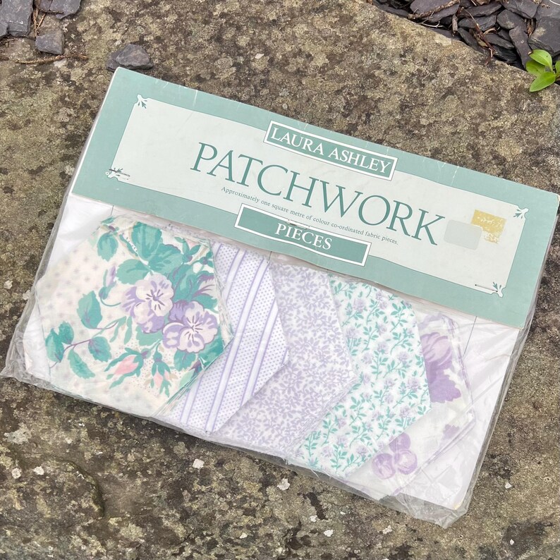 Pode incluir: Um pacote de pe&ccedil;as de patchwork Laura Ashley. O pacote cont&eacute;m v&aacute;rios peda&ccedil;os de tecido com desenhos florais e estampados em tons de branco, roxo e verde-azulado. O pacote &eacute; selado em pl&aacute;stico transparente.
