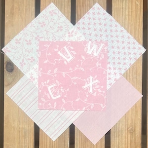 May include: A diamond arrangement of pink and white fabric squares. The central square displays the letters "V", "W", "C", and "X". The surrounding squares feature floral and striped designs. The background is a wooden surface.