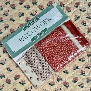 May include: A selection of Laura Ashley patchwork fabric pieces. The fabrics feature red and white floral designs. The packaging is light green and displays the words "PATCHWORK" and "PIECES". The fabrics are approximately one square metre.